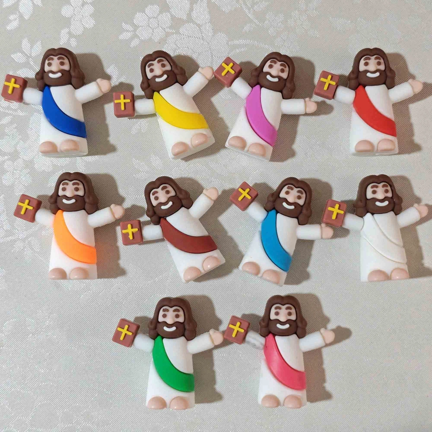 Ten colourful mini Jesus figures holding a book, arranged on lace